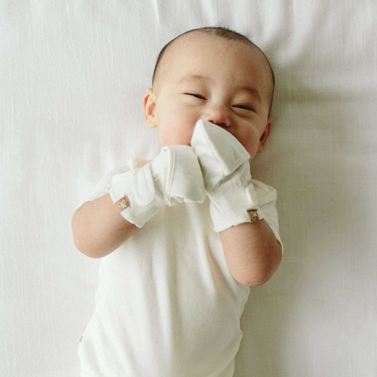 PREEMIE STAY ON MITTS in soft white fabric, worn by a baby with adjustable wrist closures and covered hands