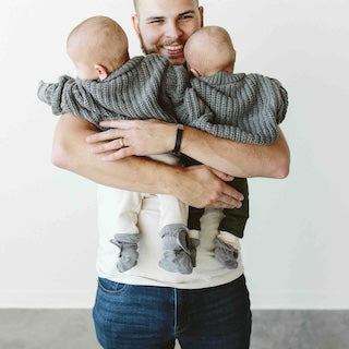 STAY ON 2-PACK BOOTS are soft grey knit baby boots with adjustable cuffs, worn by two babies in a hug