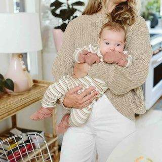 STAY ON 2-PACK MITTS are soft cream baby mittens with subtle striped cuffs, worn by a baby in a caregiver’s arms