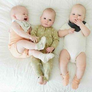 STAY ON BOOTS are soft, pale peach baby booties with adjustable straps, worn by an infant alongside two babies.