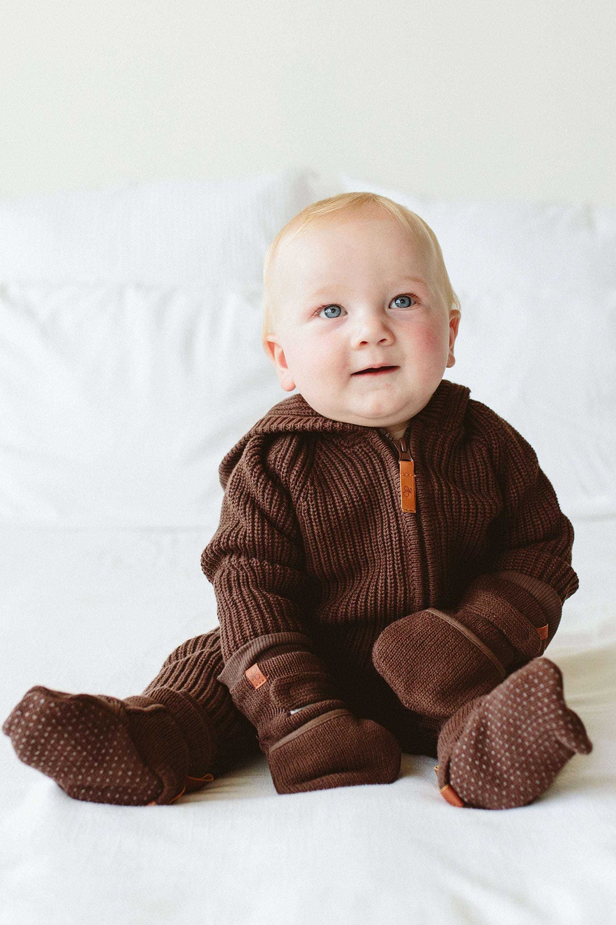 KNIT MITTS in dark brown ribbed knit with leather labels and adjustable wrist closures, worn by a baby